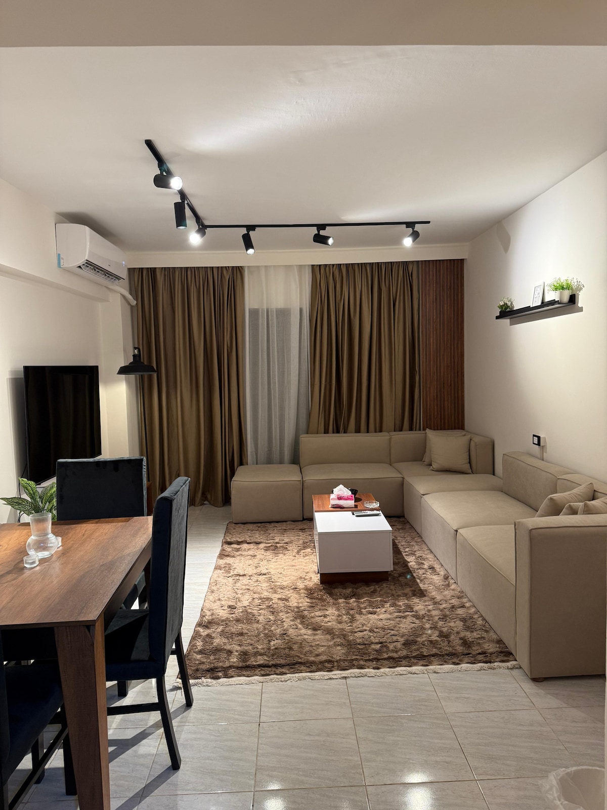 A modern living room is showcased, featuring a spacious corner couch in neutral tones and a soft area rug. Natural light is filtered through floor-length curtains. A dining table is present, accompanied by dark blue chairs, while a television is positioned against the wall.