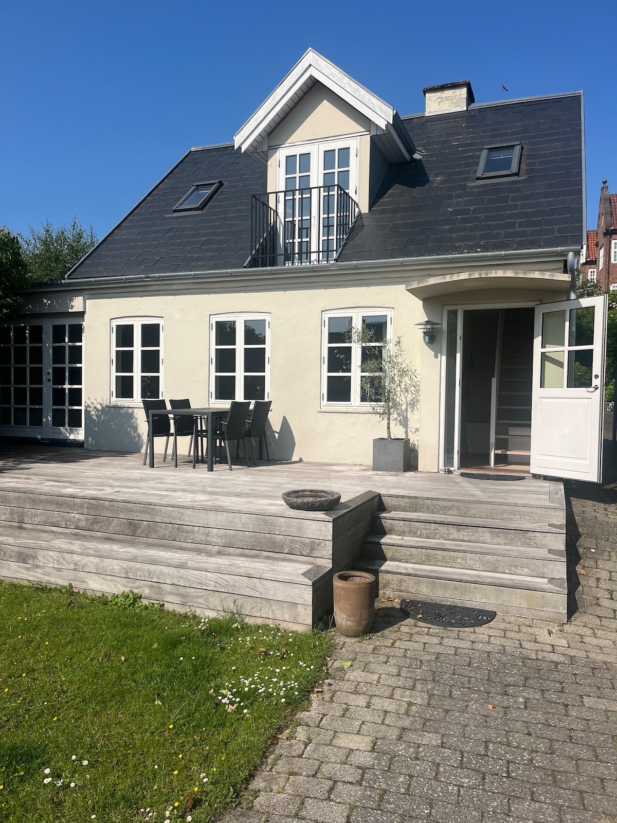 A two-story house is shown with a sloped roof and multiple windows. A wooden deck is visible in front, featuring a dining table with chairs. A grassy area is seen beside paved stones leading to the entrance, where a black doormat lays at the door.