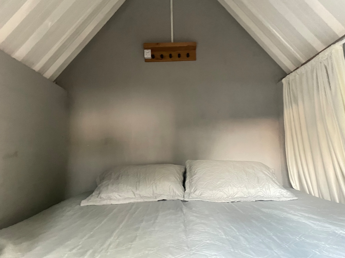 A cozy sleeping area features a spacious bed with a light-colored comforter and two matching pillows. Soft, neutral tones create a calm ambiance, while the sloped ceiling enhances the intimate feeling of the space. Curtains drape gently beside the bed, adding a touch of privacy.