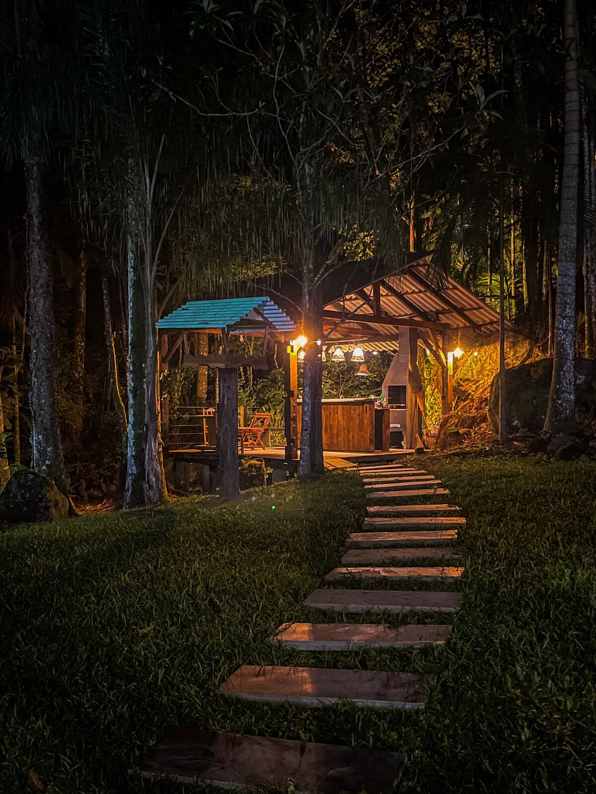 A charming rustic cabin is seen at night, softly illuminated by warm lights. A stone pathway leads through lush greenery to the cabin, which features a cozy porch area. Surrounding trees create a serene atmosphere, enhancing the connection to nature.