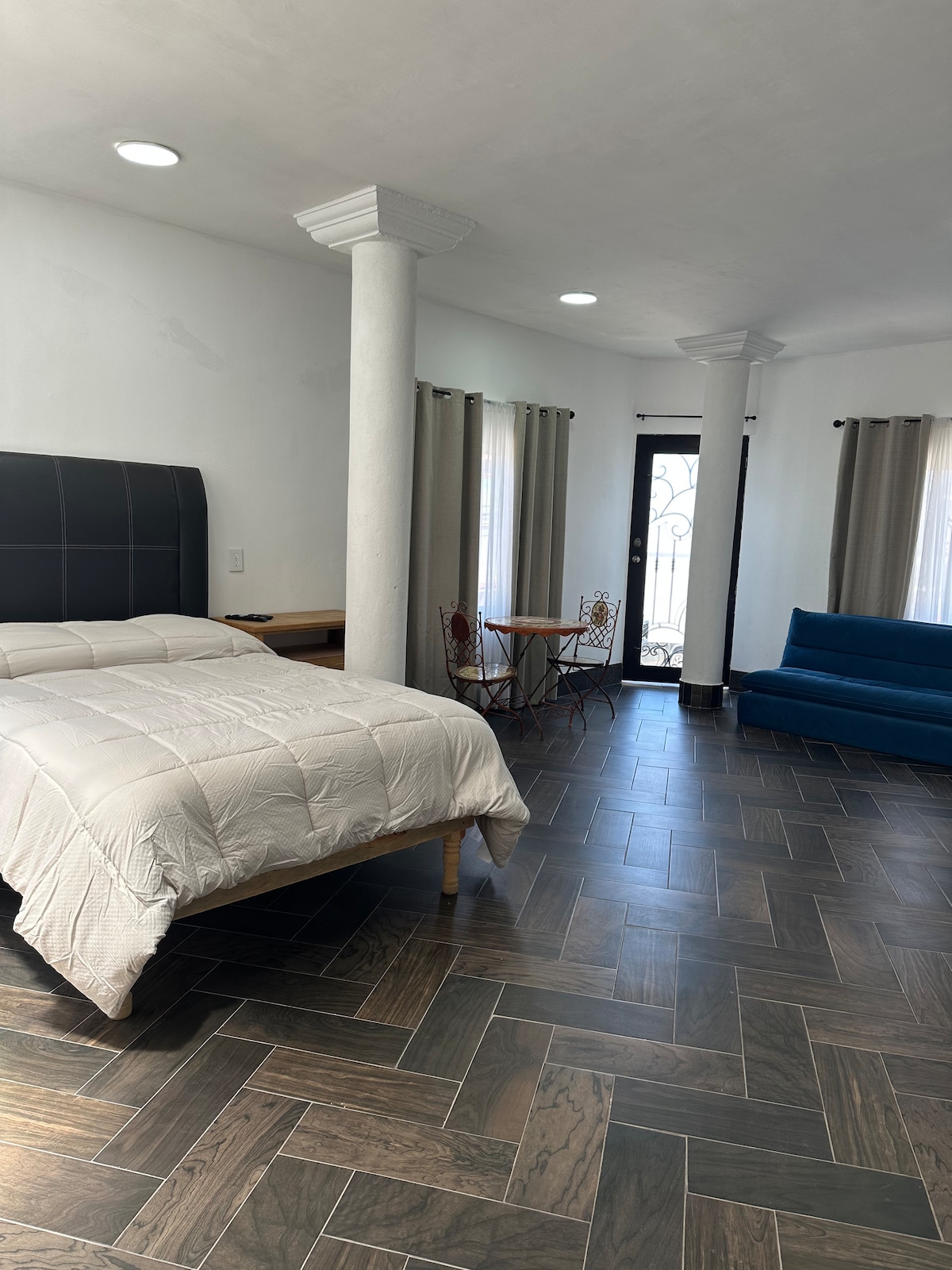 A spacious room is furnished with a large bed featuring a neutral-toned quilt. By the window, a small dining table with two chairs is set, while a comfortable blue sofa sits across the room. Natural light filters through sheer curtains, enhancing the airy atmosphere.