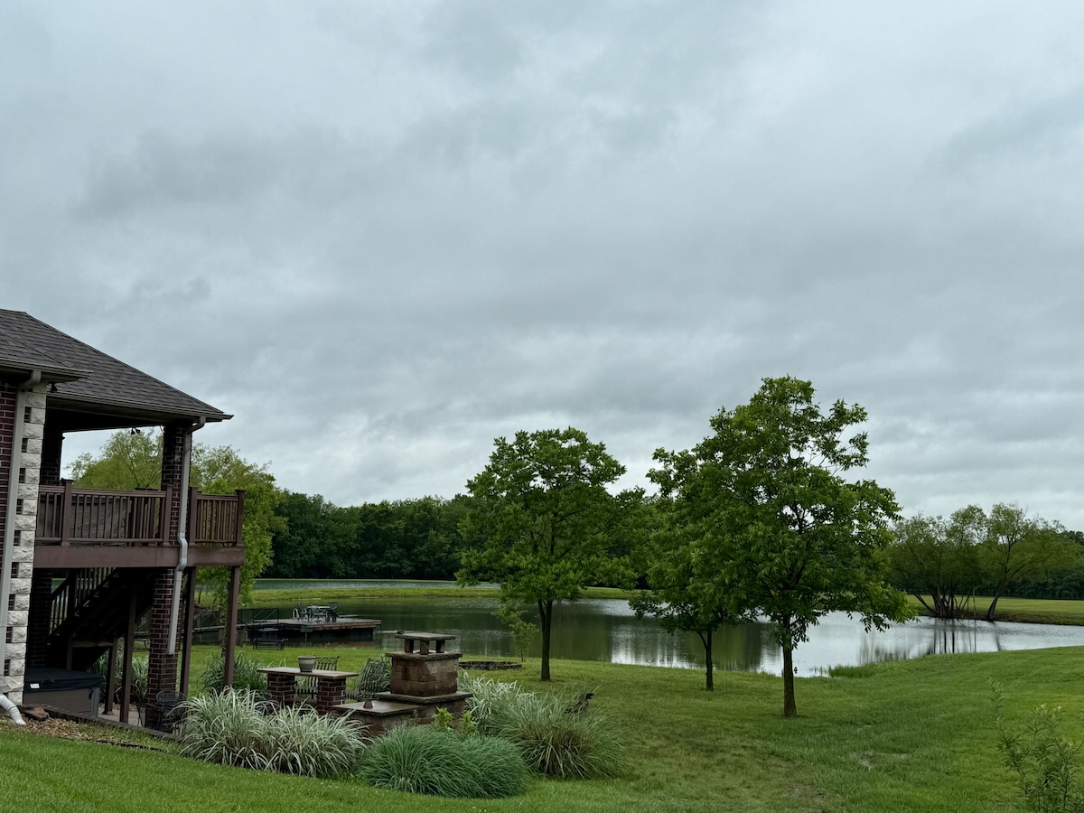 A serene view captures a backyard scene with a pond surrounded by lush greenery. A spacious deck with stairs leads to the water. Nearby, a patio area with stone seating is visible, inviting relaxation in a tranquil outdoor setting.