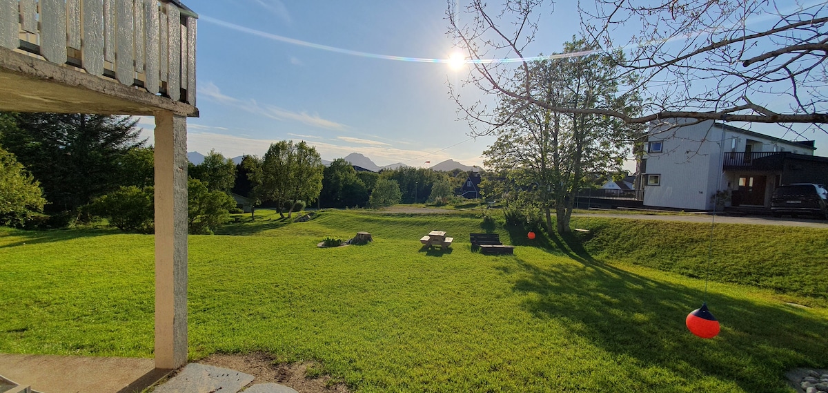 A spacious garden area is shown, featuring lush green grass under a clear blue sky. Several picnic tables are set up in a shaded section, with mountain peaks visible in the distance. The sun casts a warm light over the landscape, enhancing the peaceful ambiance.