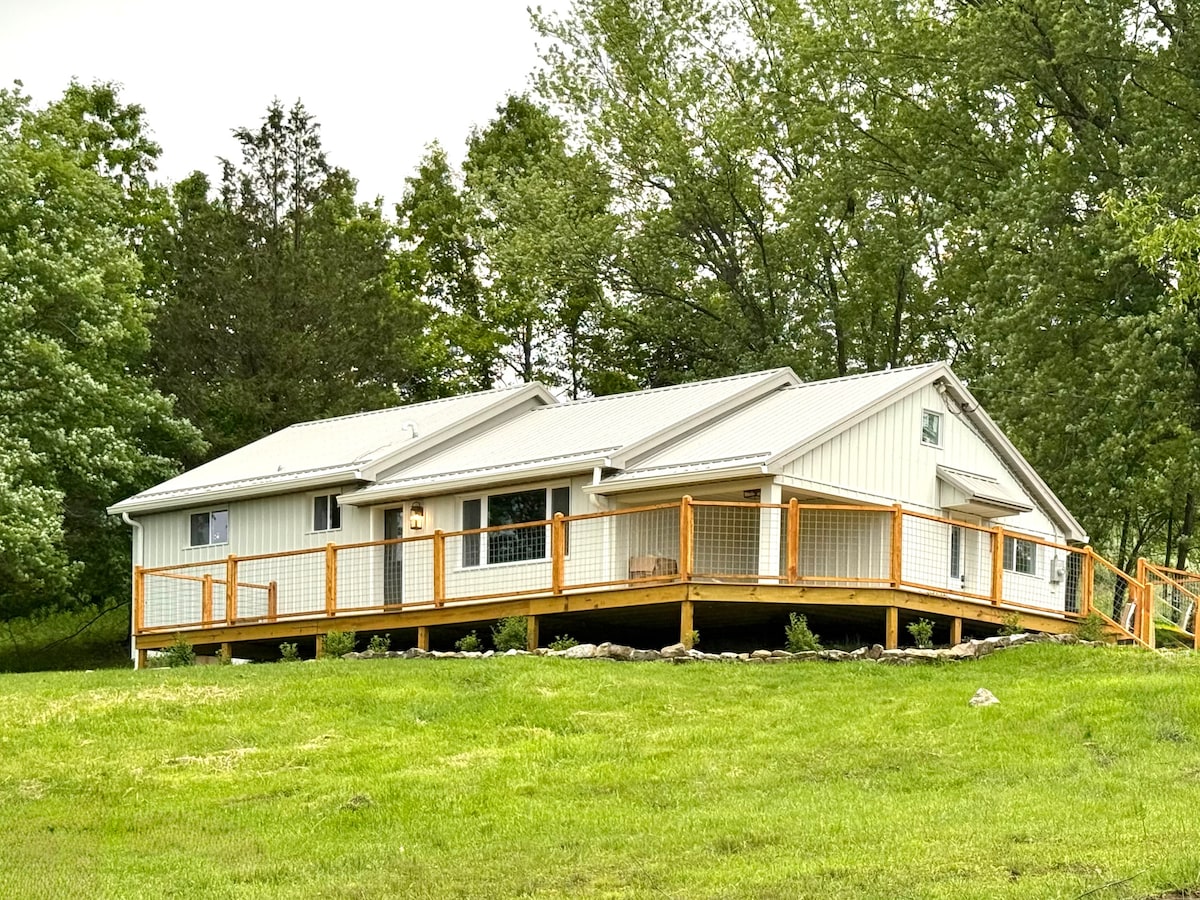 A single-story house is set amidst lush greenery, featuring a large wrap-around deck made of wood. The exterior is clad in light-colored siding, complemented by a metal roof. Windows are evenly spaced, allowing for natural light, while the surrounding landscape displays rolling hills.