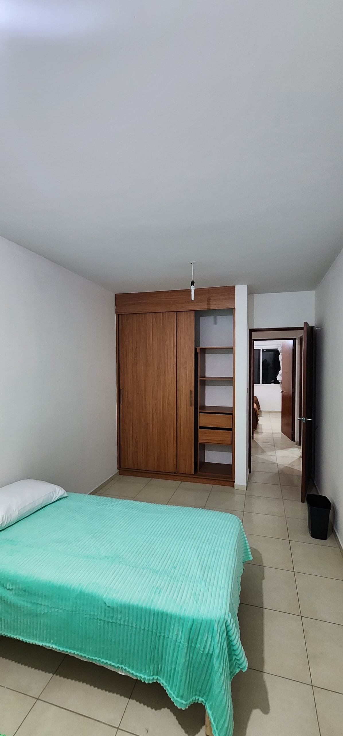 A simple bedroom features a light green bedspread draped over a spacious bed. A wooden wardrobe is embedded in the wall, displaying open shelving. The room is illuminated by overhead lighting, and a doorway leads to another area of the home.