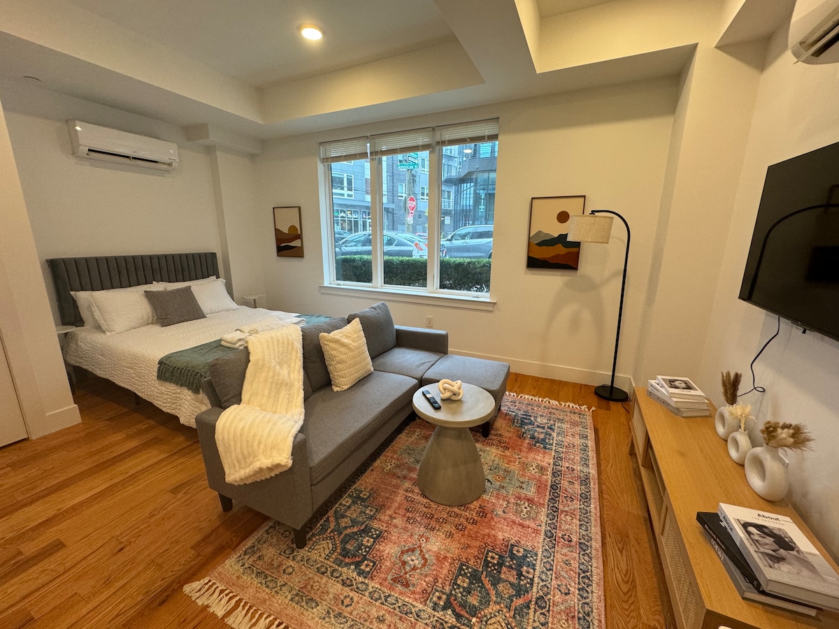 An inviting studio layout features a spacious bed with white linens, complemented by a cozy gray sectional sofa. A minimalist coffee table sits on a patterned area rug. Natural light fills the space, illuminating artwork on the walls and framing the view through large windows.