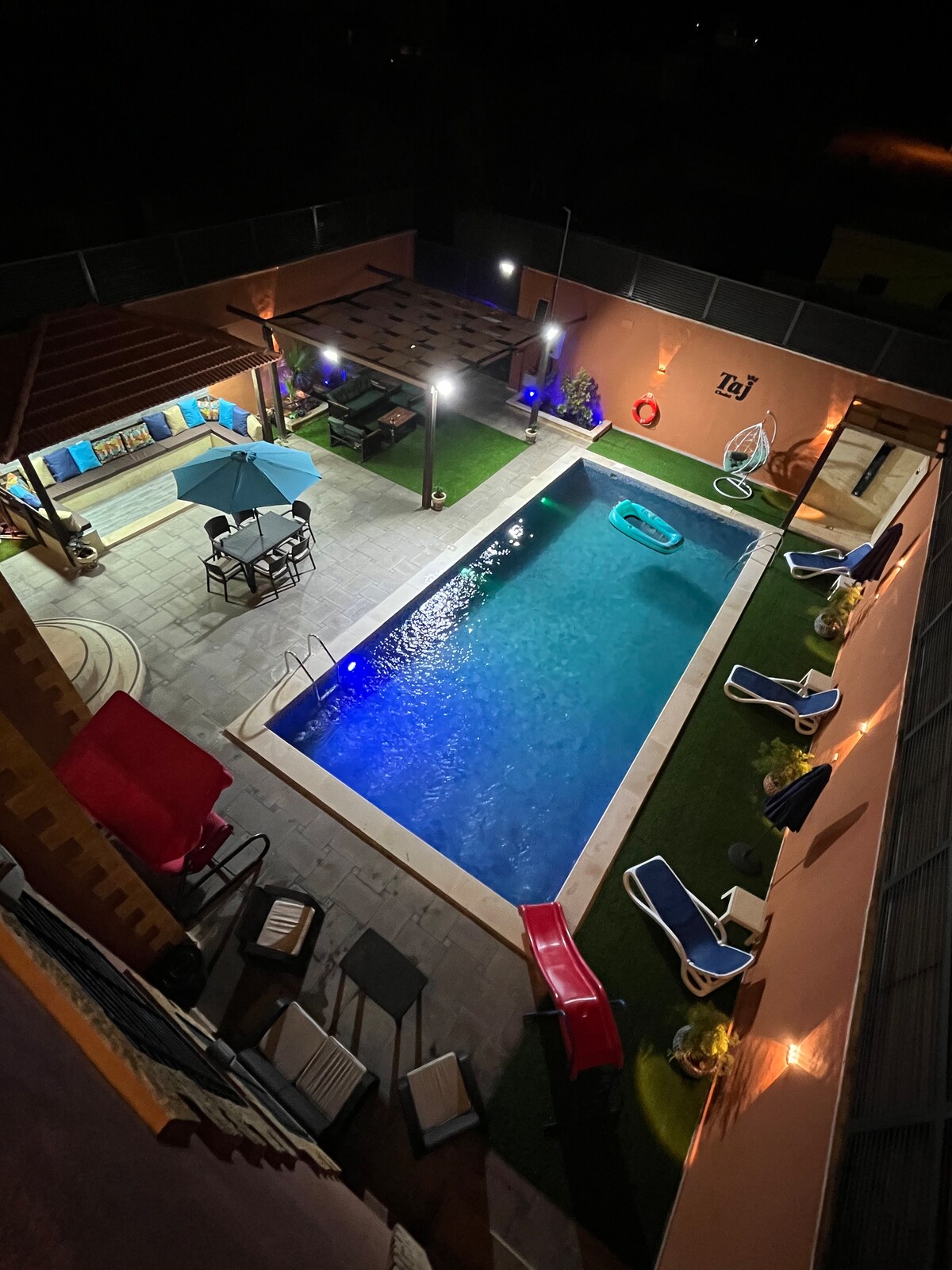 An aerial view of an outdoor space features a blue pool surrounded by relaxing lounge chairs and seating areas. Illumination enhances the pool area, while a gazebo offers additional shaded seating. Lush greenery borders the scene, creating a calm, inviting outdoor environment.
