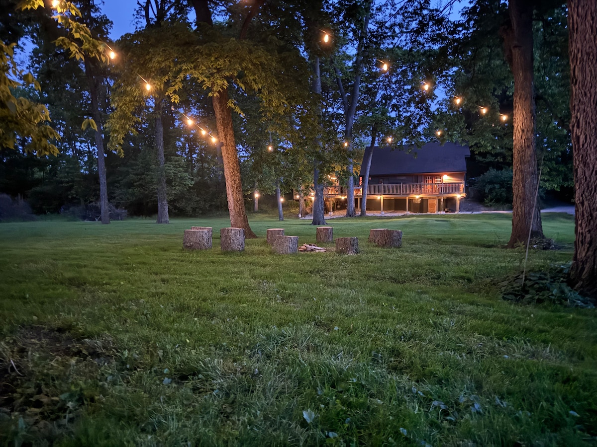 An outdoor space is illuminated by string lights, casting a warm glow over a grassy area with tree stumps arranged in a circle, suitable for gathering. In the background, a rustic cabin is nestled among trees, with soft light highlighting its façade.