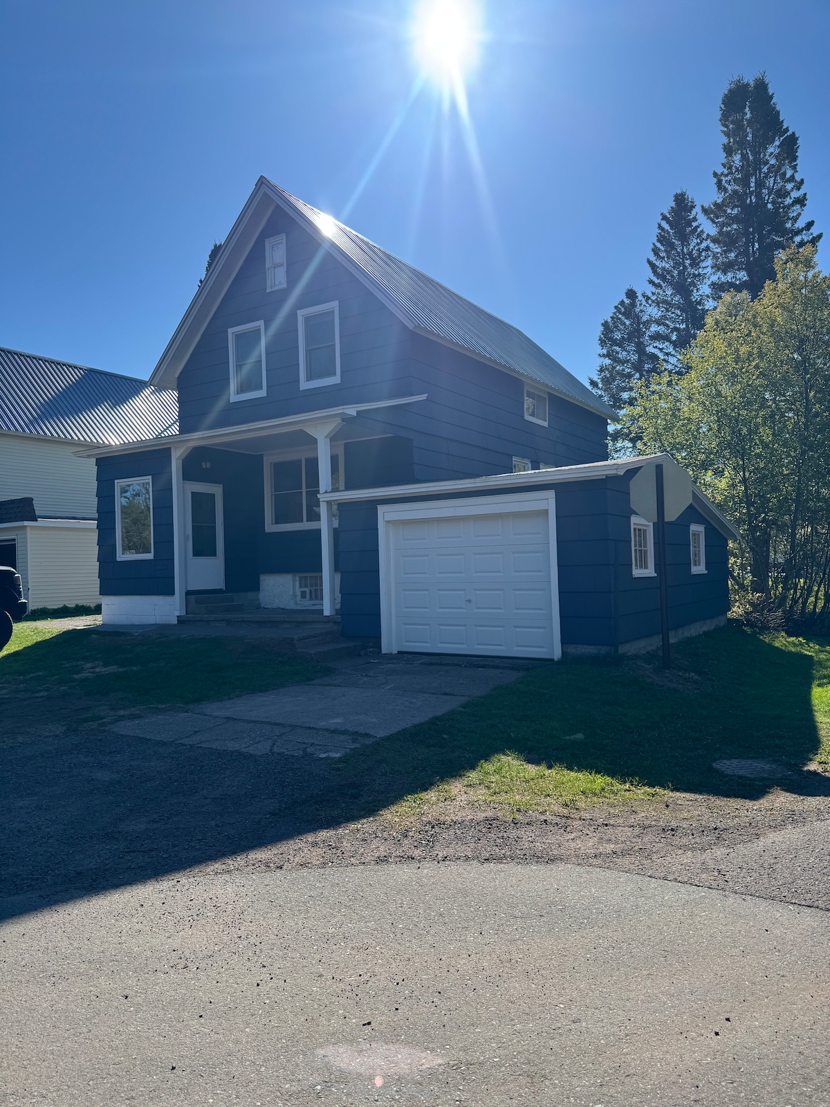 A charming blue house features a white garage, situated on a corner lot. The exterior showcases a combination of siding and a sloped roof, complemented by surrounding greenery. Sunlight casts a warm glow, highlighting the inviting setting.