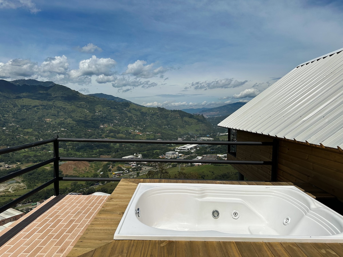 A private outdoor jacuzzi is set on a wooden deck, overlooking a mountainous landscape. The view features rolling hills and distant valleys under a blue sky, with clouds scattered across the horizon. A wooden structure is visible nearby, complementing the natural scenery.