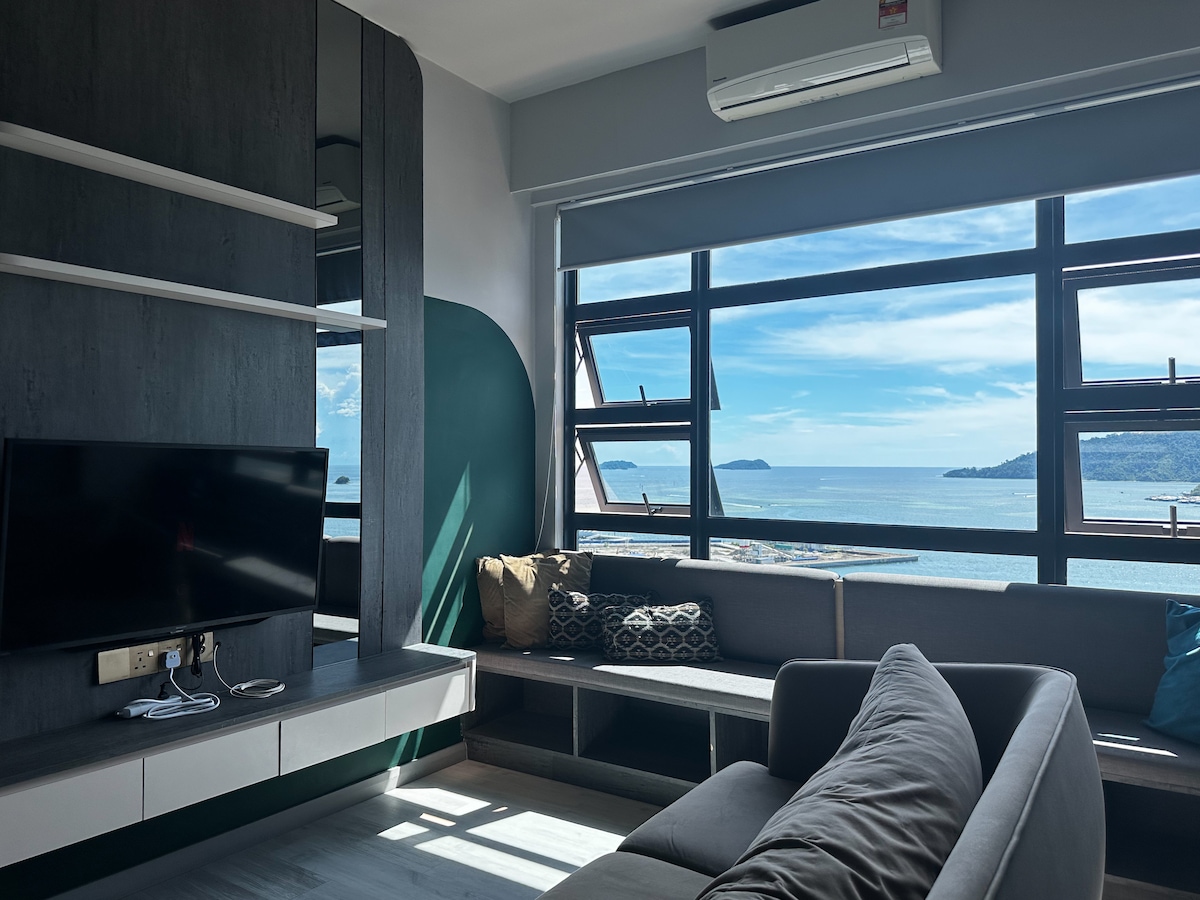 A living area is shown with large windows offering a view of the sea. A gray sofa is positioned against the wall, complemented by decorative pillows. A flat-screen TV is mounted above a minimalistic cabinet, while natural light fills the room, highlighting the clean lines of the furniture.