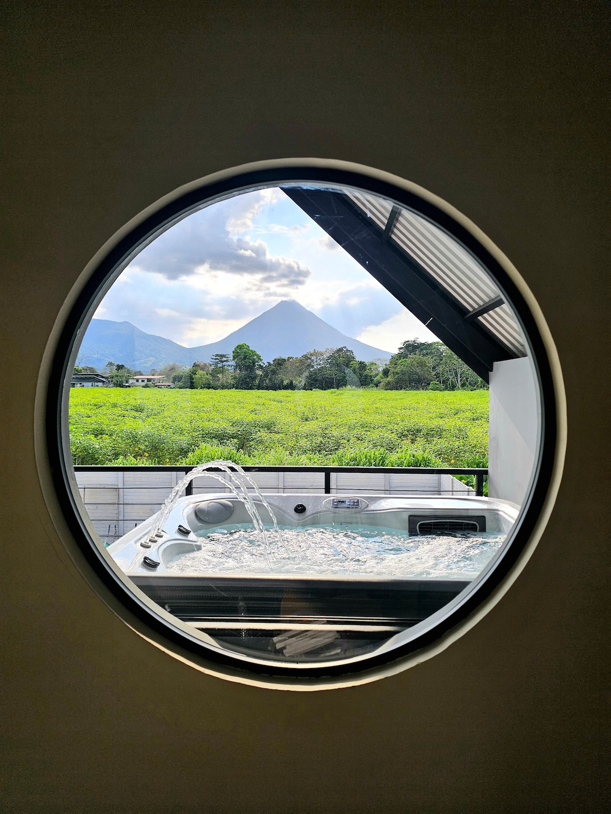 An oval window frames a view of a hydromassage jacuzzi on a private terrace, overlooking lush greenery and the silhouette of Arenal Volcano in the distance. Natural light illuminates the scene, enhancing the tranquil atmosphere of the surrounding landscape.