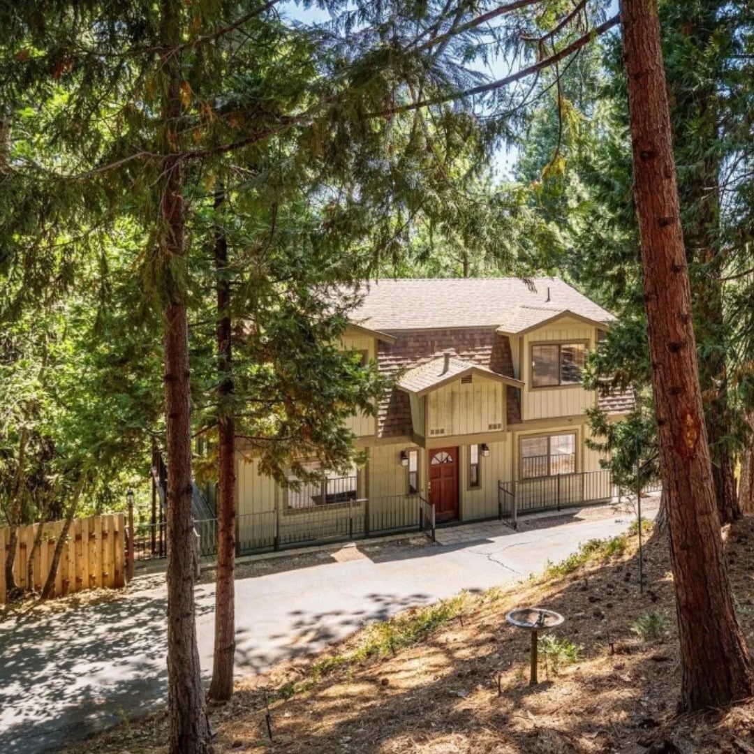 A two-story house is set amidst tall trees, featuring an inviting facade with large windows and a central entrance. A gated pathway leads to the door, providing a sense of privacy. Sunlight filters through the surrounding greenery, creating a peaceful ambiance.