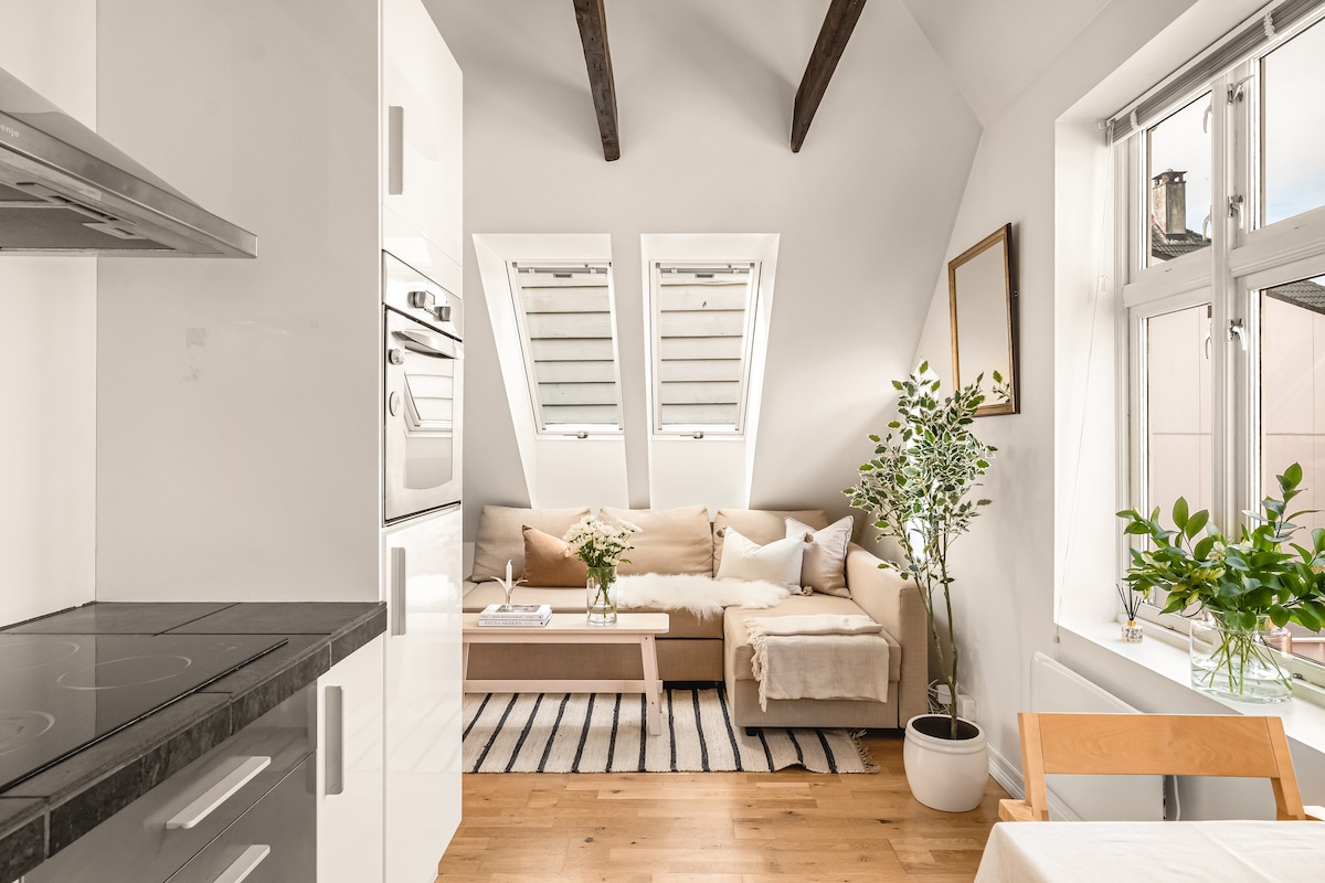 A bright living area is featured with a light-toned sofa adorned with soft throw blankets. The room includes an adjacent coffee table and a potted plant. Natural light filters through triangular windows alongside a framed mirror, enhancing the airy atmosphere of the space.