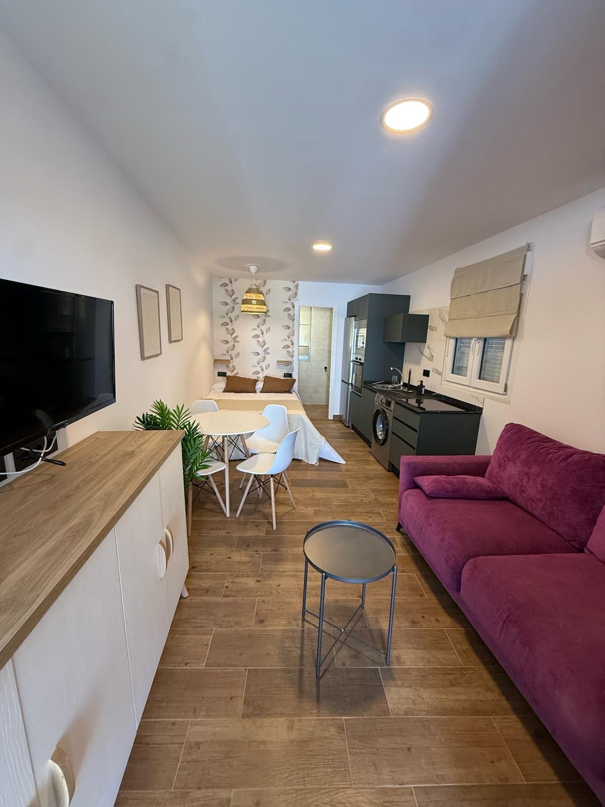 A modern living space features a purple sofa, a wooden entertainment unit holding a television, and a light-colored dining area with white chairs. The kitchenette includes dark cabinetry and appliances, while soft lighting enhances the atmosphere. Neutral wallpaper with a subtle pattern adds a decorative touch.