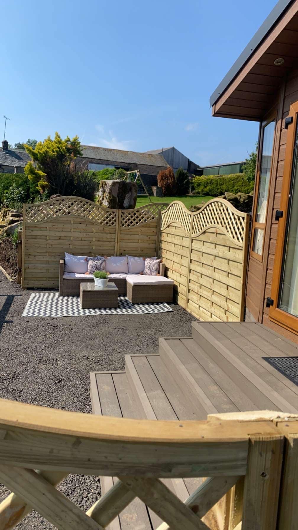 A seating area is positioned outside the caravan, featuring a small sofa with cushions set against a wooden fence. The space is complemented by a patterned rug on the gravel surface, surrounded by greenery and a clear blue sky.