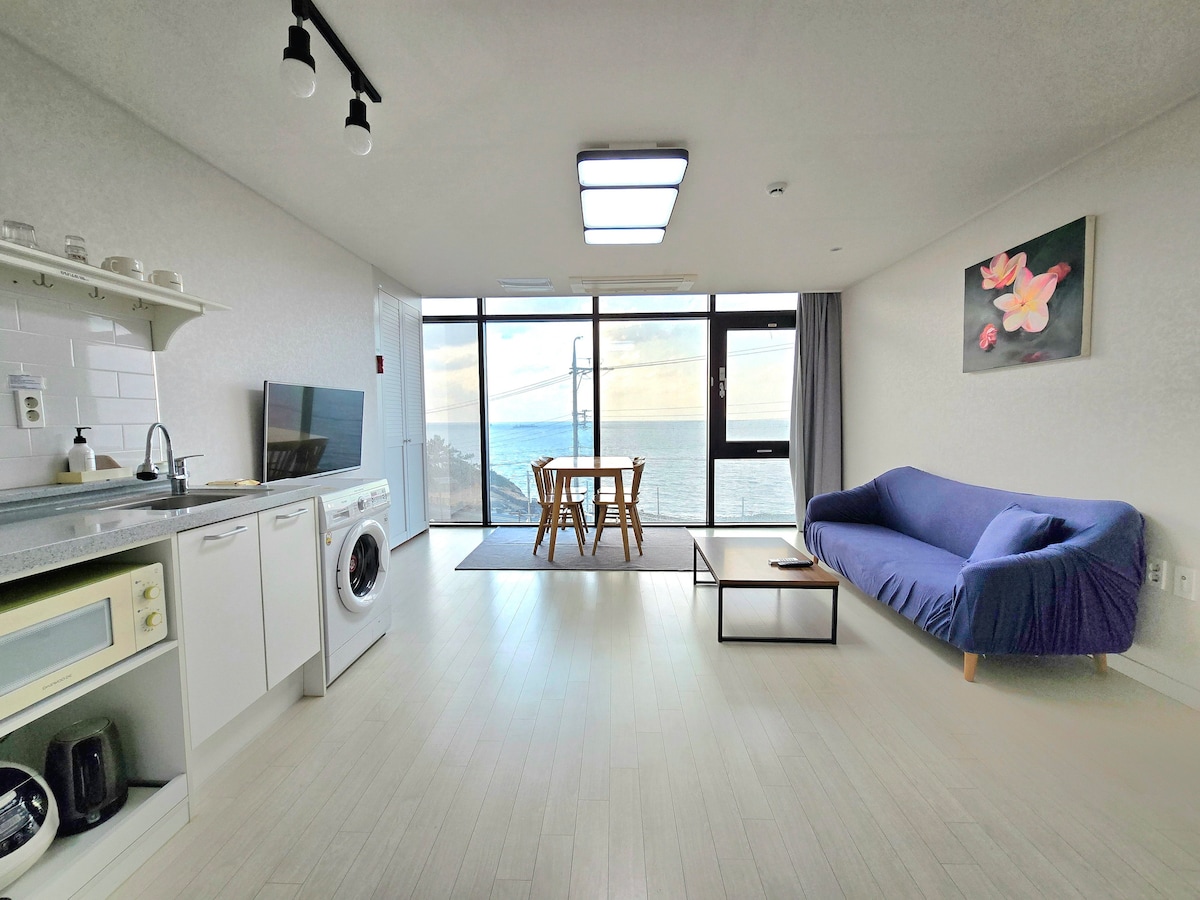 An open living space features large windows offering ocean views. A cozy blue sofa is positioned opposite a dining table set for four. The kitchen area includes essential appliances, while a washing machine is conveniently placed nearby. Soft natural light fills the room.