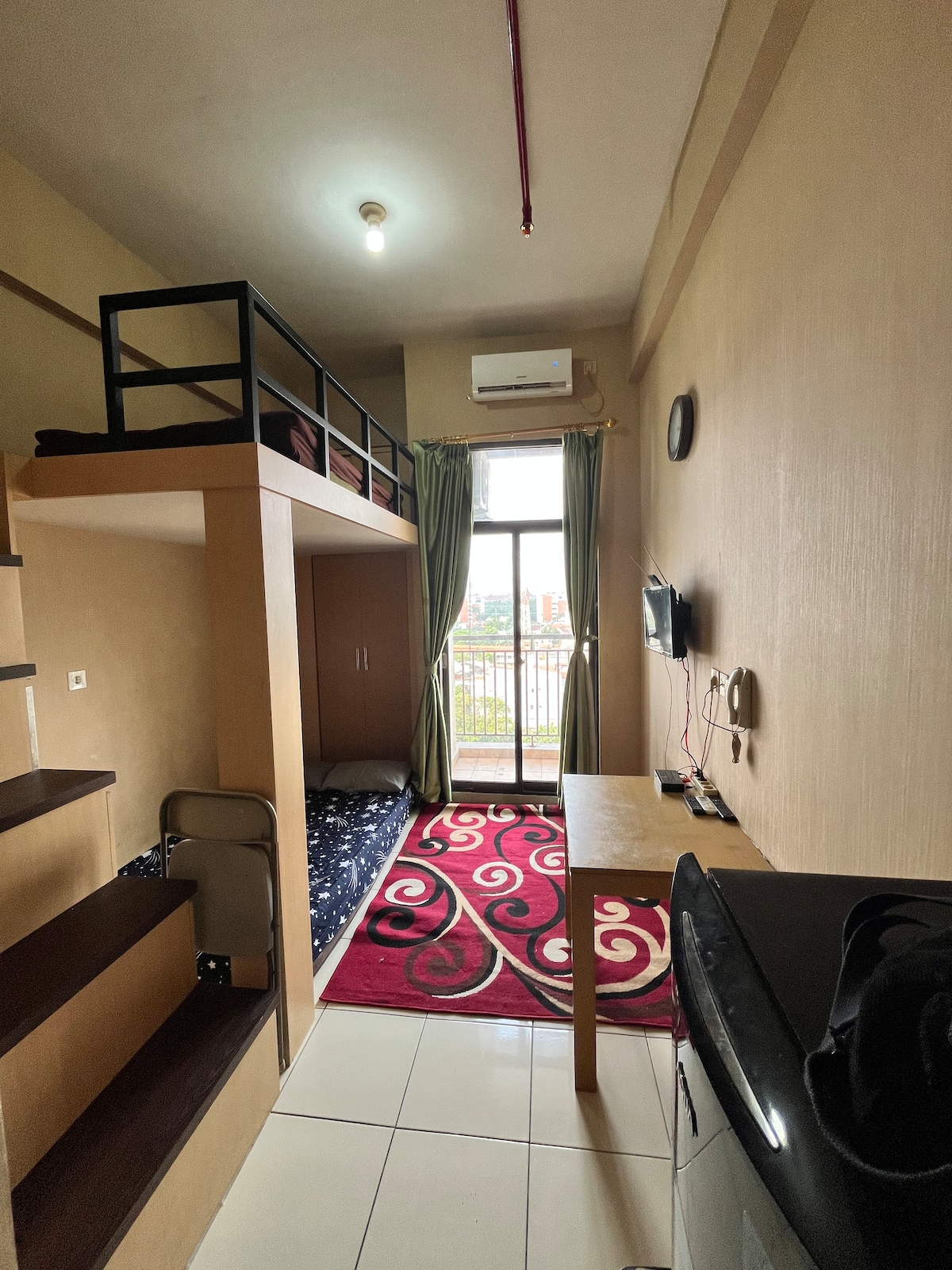 A compact room features a lofted sleeping area accessible by stairs. Below, a cozy space is available for additional activities. A television is mounted on the wall opposite the large window, which is dressed in light curtains, allowing natural light to fill the room. A vibrant area rug adds color to the tiled floor.