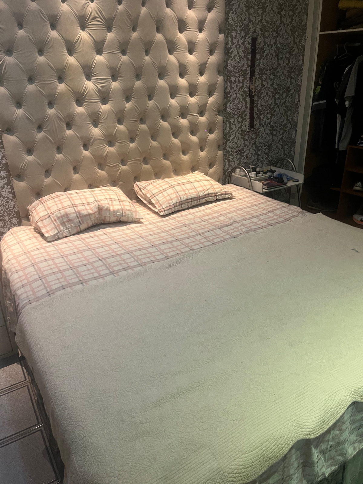A spacious bed with a tufted headboard is presented, featuring two decorative pillows and a soft blanket draped across the top. In the background, a closet is visible, displaying hangers and a few folded items, contributing to the room's organized appearance.