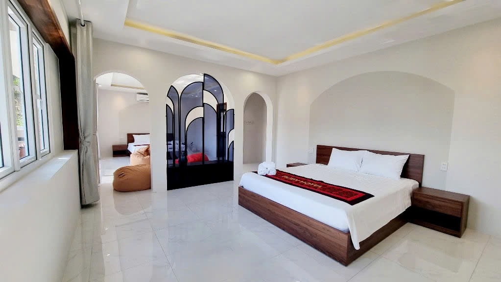 A spacious bedroom features a large bed with a neutral-colored bedspread and dark wood furniture. A unique room divider adds depth to the layout, while natural light floods through multiple windows, illuminating the minimalist decor.