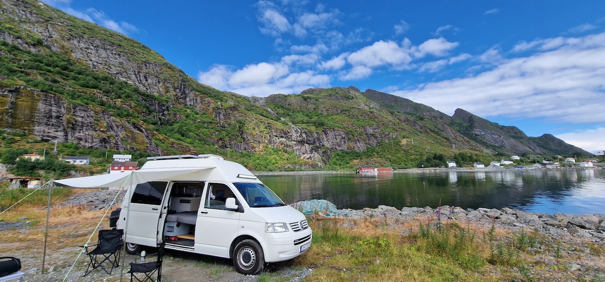 A white Volkswagen camper van is parked on a grassy area by a tranquil body of water, surrounded by towering green mountains. Two black camping chairs are set up alongside the van, and a white awning provides shade over the entry. The scene captures a serene, natural landscape.