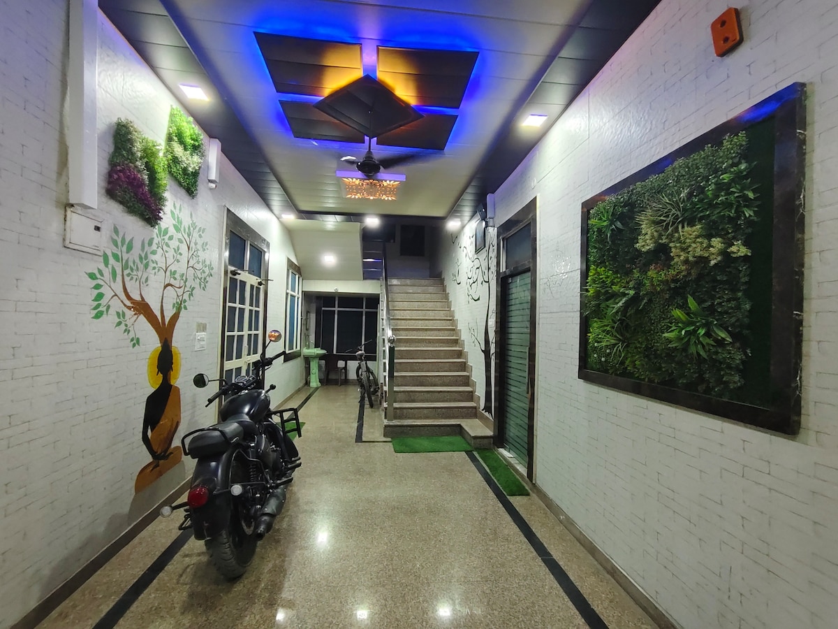 A well-lit entrance features a modern ceiling with colorful lighting above a staircase. A black motorcycle and bicycle are parked near the wall. Greenery is displayed in wall art and a large frame, while the white brick walls are adorned with decorative tree motifs.
