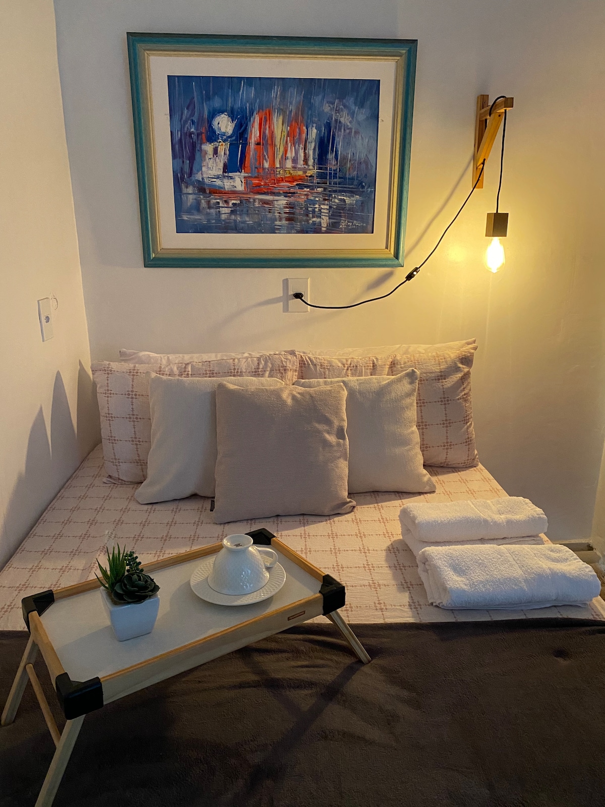 A cozy sleeping nook is featured, furnished with a low mattress topped with decorative pillows. A small side table holds a teapot and cup, while fresh towels are neatly arranged nearby. The wall is adorned with a colorful artwork, and warm light highlights the space.