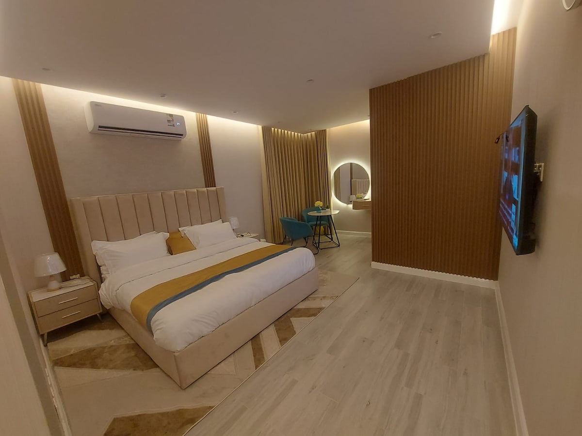 A modern bedroom features a spacious bed with a simple headboard and colorful bedding. A small table and chair are positioned near a round mirror, while a television is mounted on the wall. Soft lighting is provided by accent walls and ceiling fixtures.
