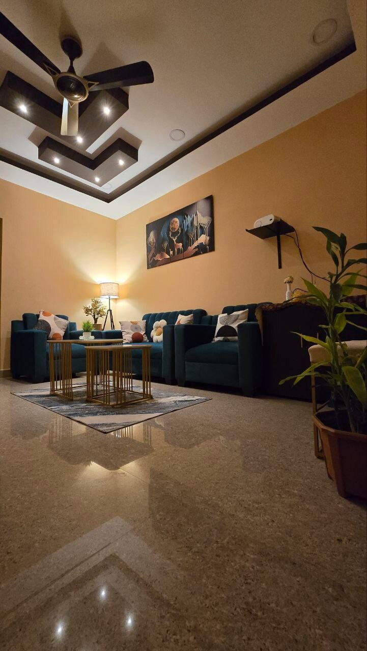 A living area is portrayed with teal sofas arranged around a coffee table. Soft lighting is provided by a floor lamp, complementing the light-colored walls and decorative art piece above. A potted plant adds a touch of greenery, while polished flooring reflects the space.