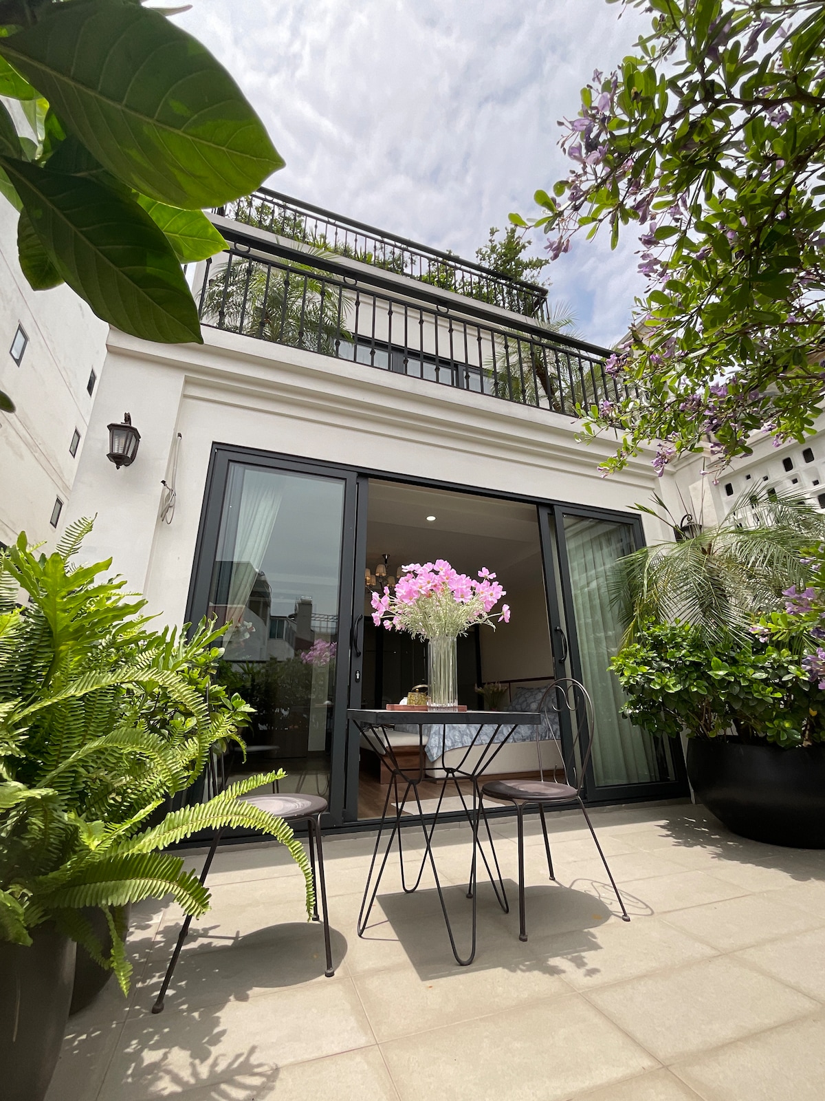 A private outdoor seating area is framed by lush greenery, featuring a small round table and two chairs. A vase filled with pink flowers is positioned at the center of the table. Large glass doors provide access to the interior, enhancing the airy ambiance.