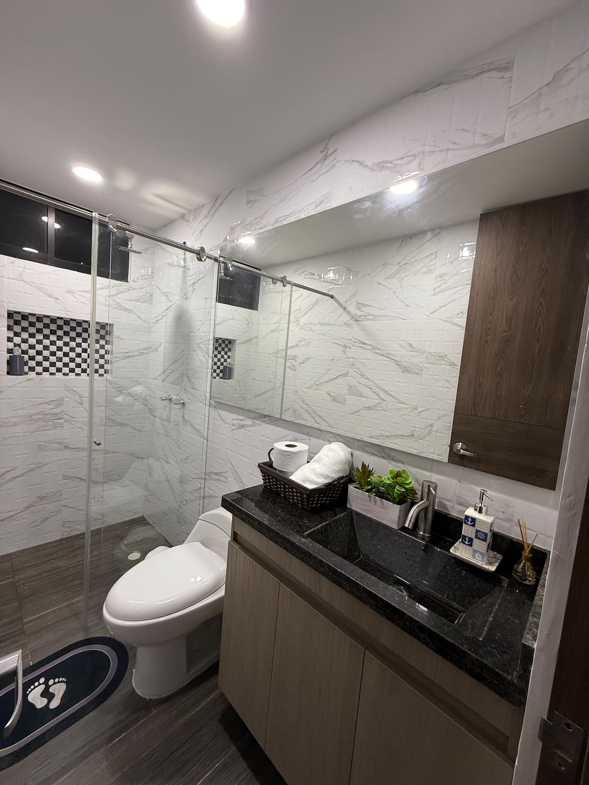 A modern bathroom features white marble walls, a glass shower enclosure, and a dark granite vanity. Light fixtures illuminate the space, while a woven basket holds rolled towels. A decorative plant adds a touch of greenery, and a bath mat with a footprint design is positioned in front of the toilet.