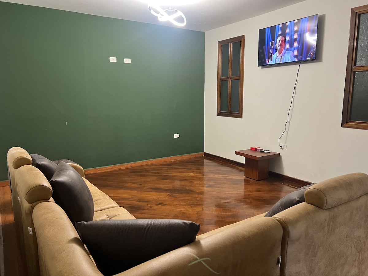 A spacious living area features a comfortable beige sectional sofa with dark pillows. A flat-screen television is mounted on the wall across from the seating. Large windows allow natural light to fill the room, which is accentuated by a deep green wall.