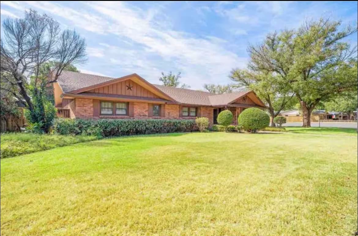 A well-maintained exterior of a single-story home is presented, featuring a brick facade and a sloped roof. A spacious lawn surrounds the property, showcasing trimmed hedges and scattered trees. The house is set in a quiet neighborhood, with a clear blue sky above.