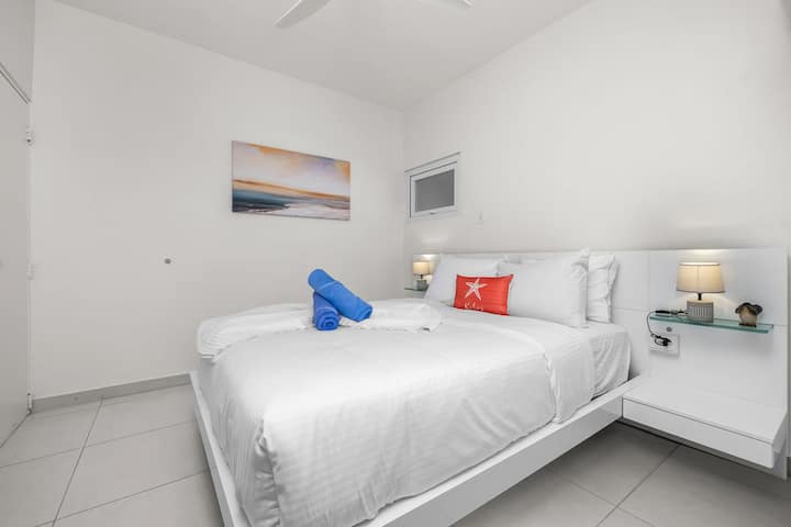 Dormez profondément dans vos draps d'un blanc éclatant, entourés de tables de chevet flottantes modernes et d'un éclairage doux au chevet. Une œuvre d'art côtière sereine crée une ambiance tranquille parfaite.