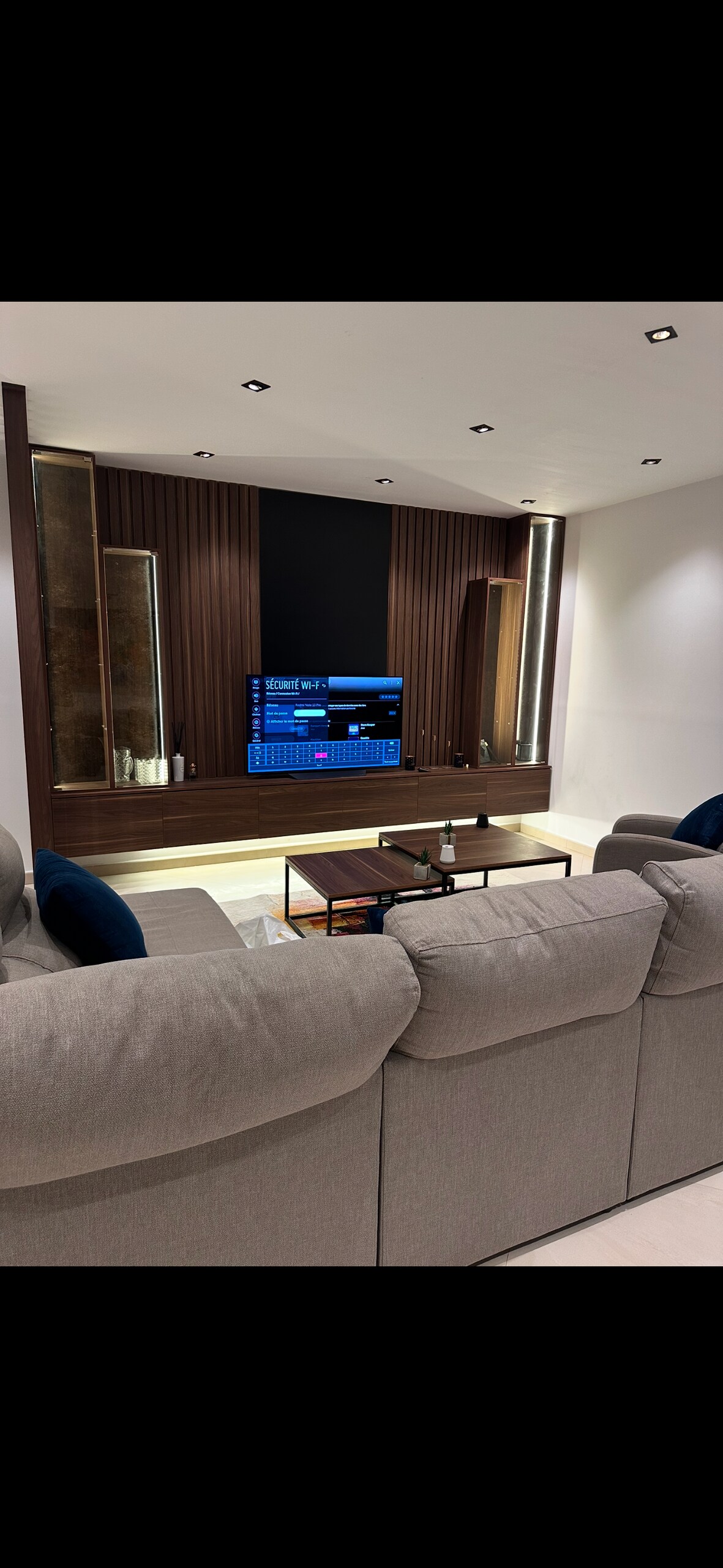 A modern living room is presented with a comfortable gray sofa facing a sleek entertainment unit. The unit features a flat-screen TV mounted on a dark wall, complemented by accent lighting. A low coffee table sits in front, adding functionality to the space.