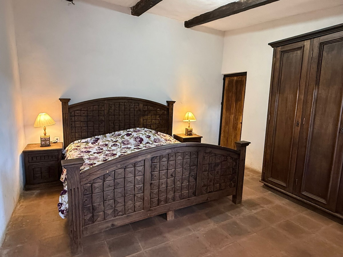 A spacious sleeping area is highlighted by a large wooden bed adorned with a floral patterned spread. Two bedside lamps provide soft illumination. A sturdy wooden wardrobe and a small door are visible, suggesting ample storage space.