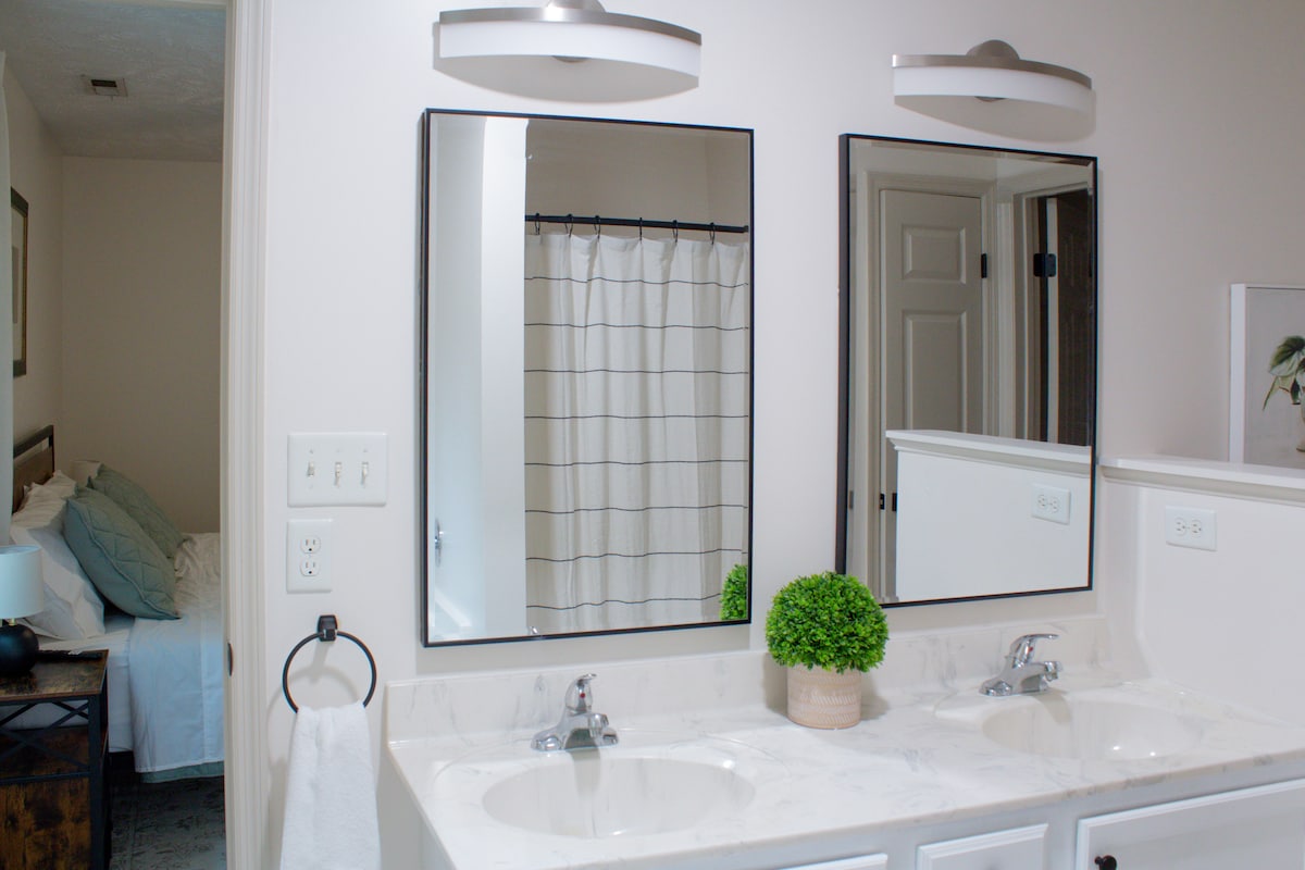 A modern bathroom is shown with dual sinks under two sleek mirrors. A light-colored countertop contrasts against the white cabinetry. A shower curtain with a grid pattern is visible in the background, and a small potted plant adds a touch of greenery to the space.