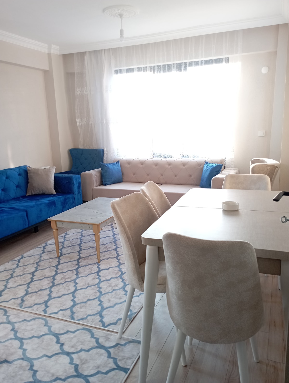 A bright living space features a beige sofa and two blue armchairs, creating a comfortable seating area. A light-colored table is placed in front of the sofa. A dining area with beige chairs is adjacent to a window, allowing natural light to fill the room.