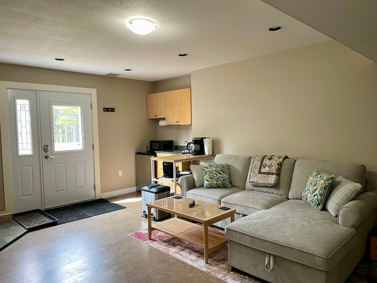 A cozy living area is presented with a spacious sectional sofa adorned with decorative cushions. A wooden coffee table rests in front of the sofa. The kitchenette, equipped with appliances, is visible in the background, alongside a welcoming entrance area featuring a front door and a small rug.