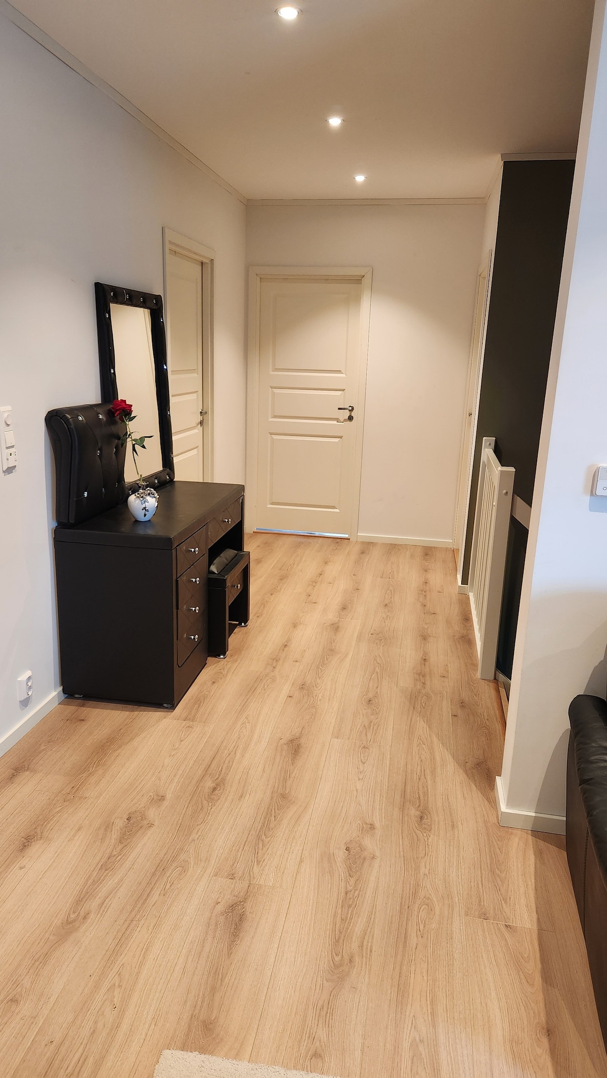 A well-lit hallway features a smooth wooden floor and light-colored walls. A black console table with a mirror and decorative items is positioned against one wall. Two doors lead to additional rooms, while a dark accent wall adds depth to the inviting space.