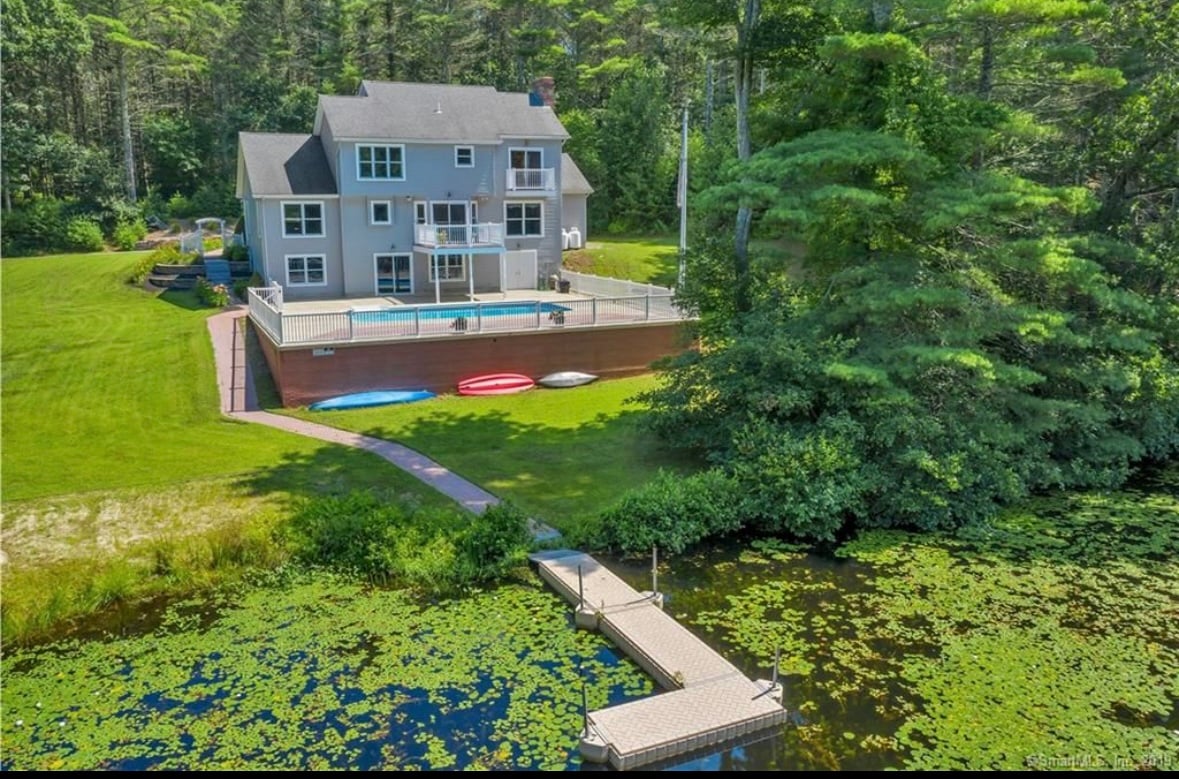 The image captures a spacious backyard featuring an in-ground pool surrounded by a wooden deck. Several kayaks are lined up on the grass, with a dock extending toward the pond, framed by lush greenery and tall trees in the background.