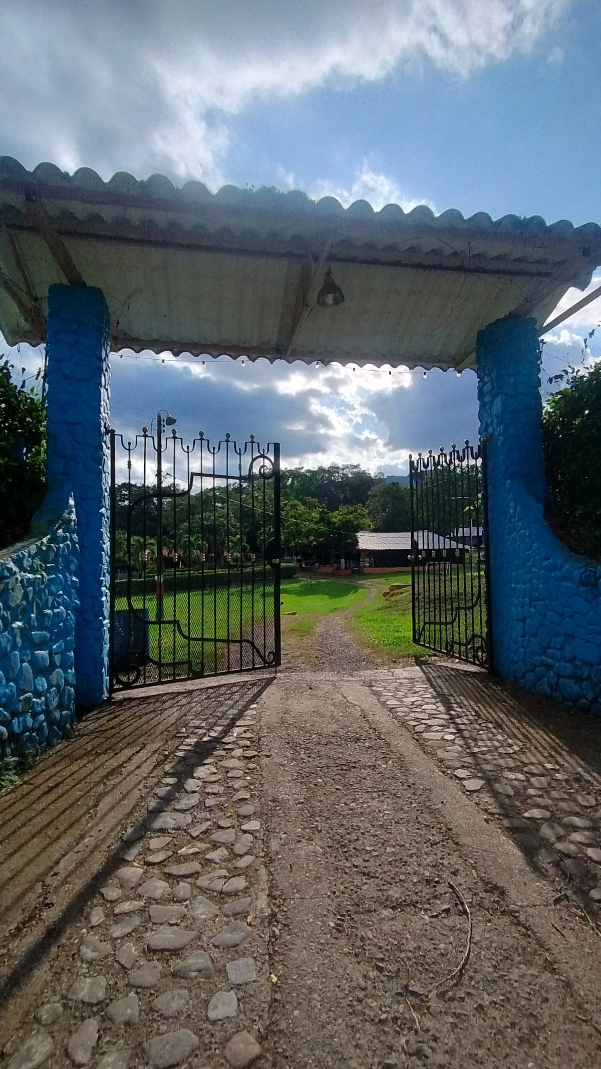 An open blue gate leads into a green landscape, framed by stone walls. A gravel path can be seen extending into the distance, with natural light illuminating the scene under a partly cloudy sky.