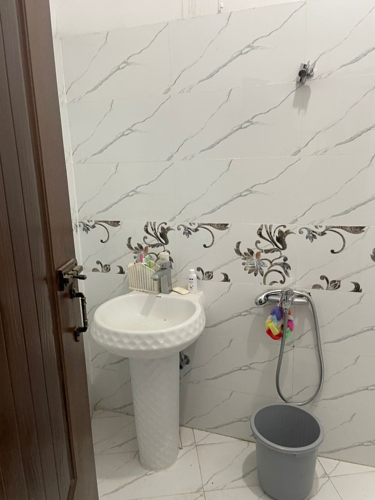 A neatly arranged bathroom is equipped with a white pedestal sink and chrome fixtures. Decorative tiles with floral patterns enhance the space. Personal care items are organized on a shelf, and a small waste bin is positioned nearby, contributing to the bathroom's functionality.