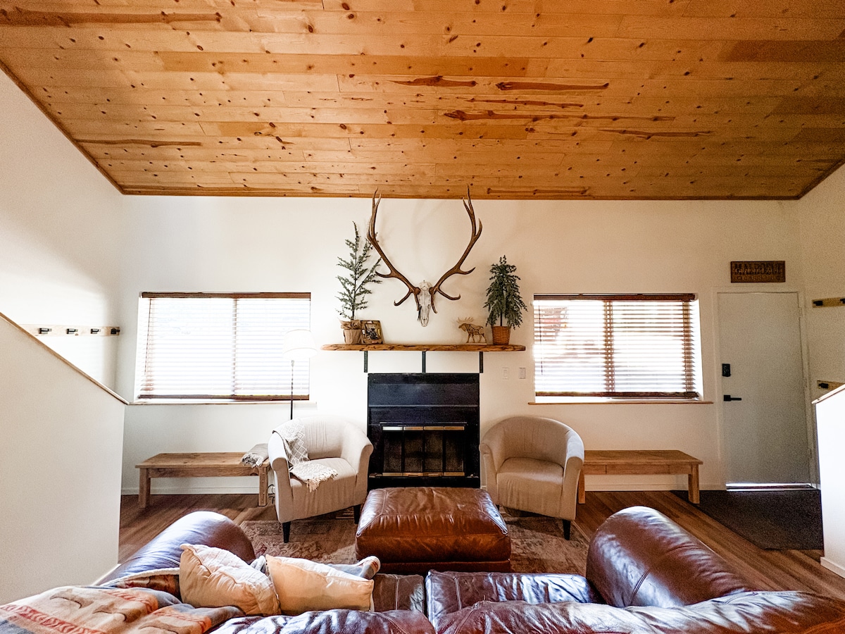 A spacious living area features a leather sofa and armchairs, paired with a wooden coffee table. Natural light enters through two windows adorned with blinds. An antler decoration is mounted above the fireplace, flanked by potted plants. A welcoming atmosphere is created by the wooden ceiling.