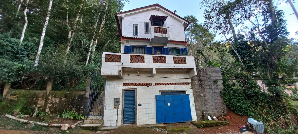 A three-story house is set among trees, featuring a white exterior with blue shutters and a tiled roof. A terrace with decorative railings is visible on the second level, while the ground level showcases a blue garage door. Lush greenery surrounds the property.