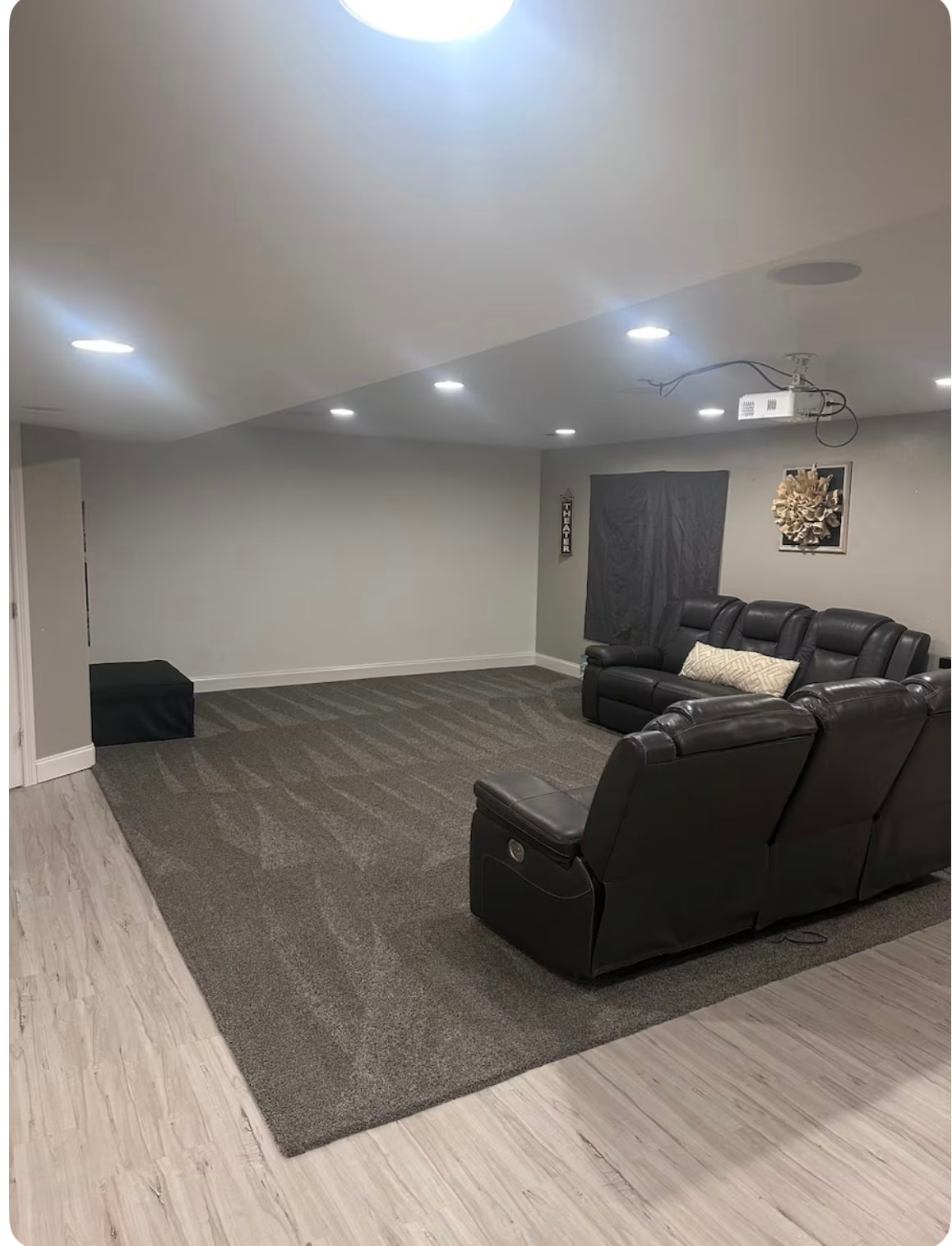 A spacious living area is shown, featuring plush black leather recliners arranged for relaxation. Soft gray carpeting covers the floor, while overhead lighting illuminates the room. A dark panel covers a small doorway, adding a touch of privacy to the decor.