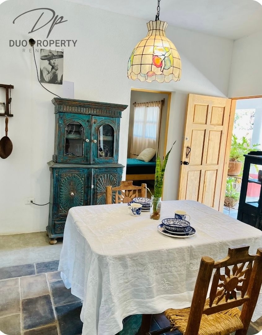 A bright dining area features a rustic wooden table covered with a white tablecloth. Two blue and white plates are set at each place. An ornate cabinet stands against the wall, and a colorful pendant light provides soft illumination.