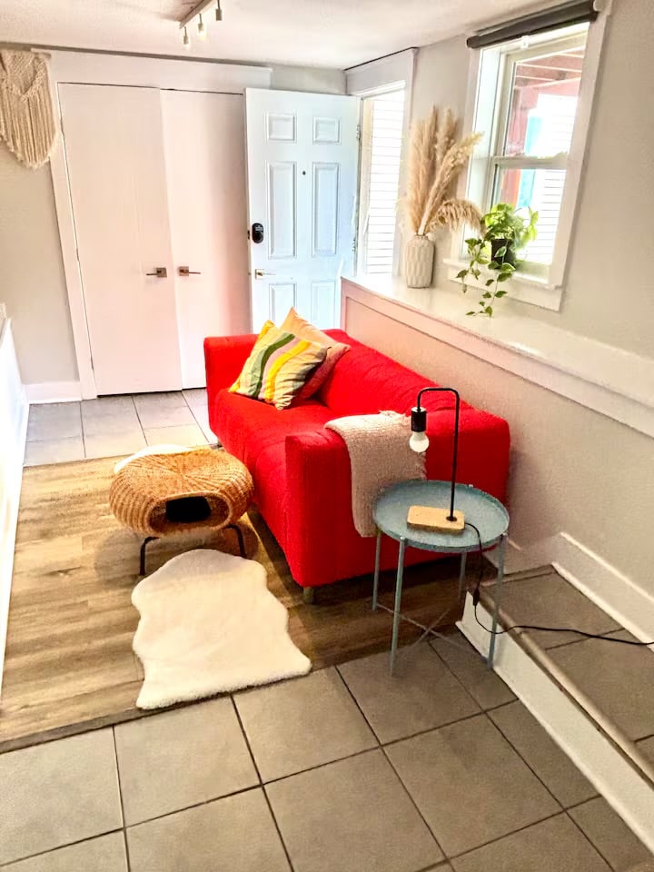 A cozy corner features a bright red sofa adorned with colorful accent pillows, a small blue side table with a lamp, and a woven stool. Natural light enters through a nearby window, illuminating the tiled floor and creating a warm atmosphere.