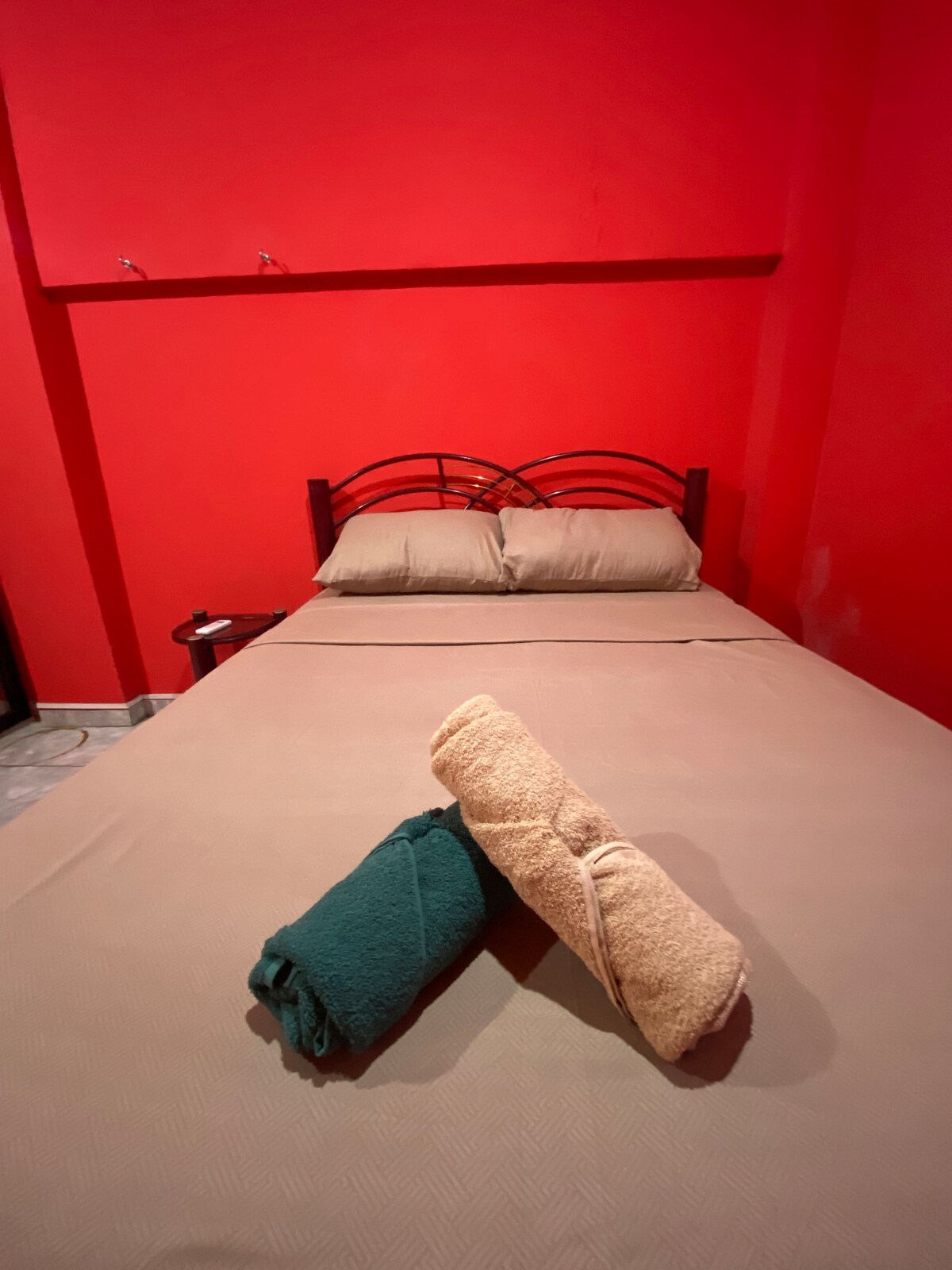 A cozy sleeping area features a spacious bed with neutral-colored linens against a vibrant red wall. Two neatly rolled towels in contrasting colors are placed on the bed, adding a touch of comfort to the space.