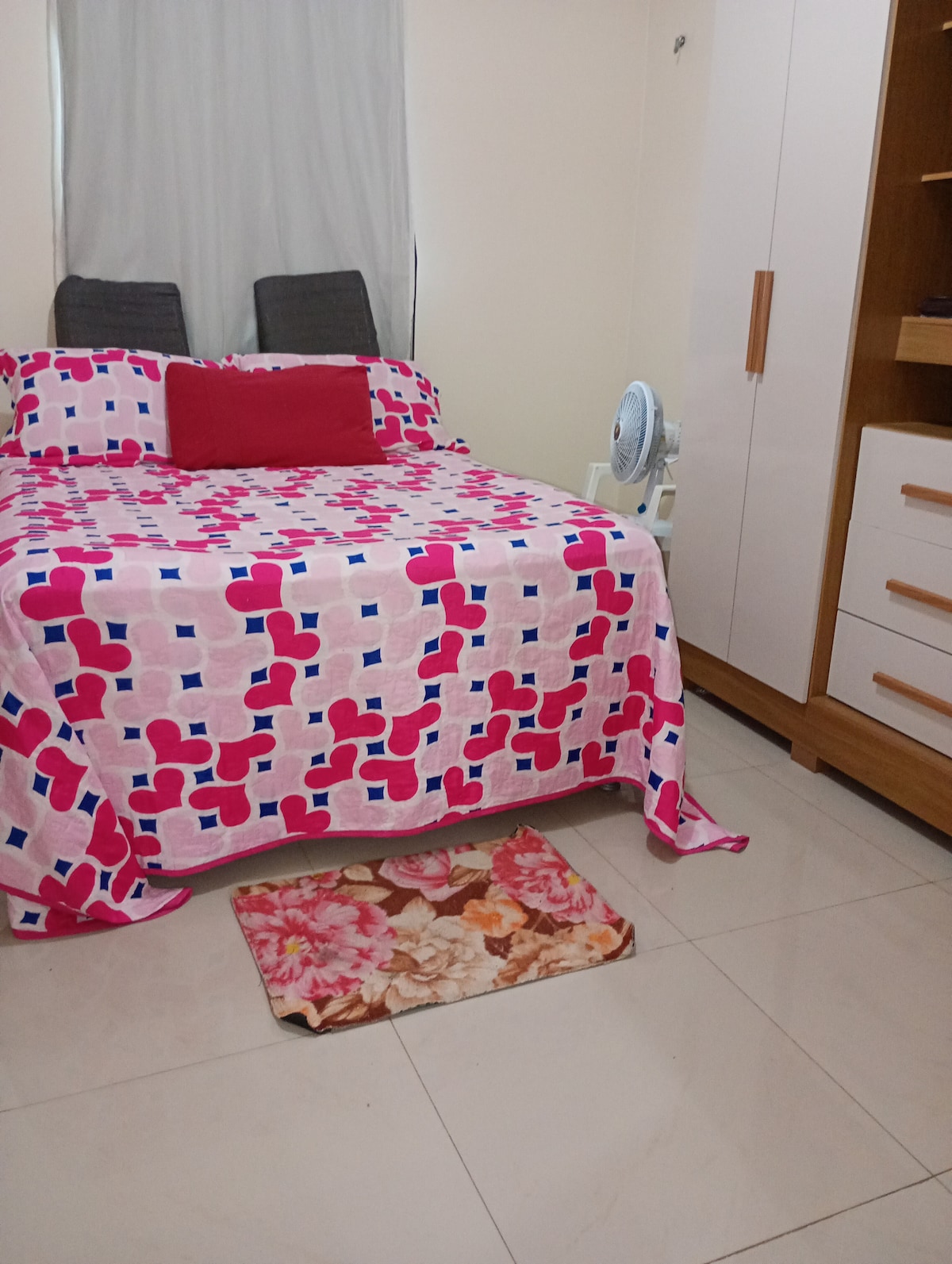 A simple sleeping area is furnished with a double bed covered in a colorful patterned blanket. Two upright chairs are positioned at the back. A small fan provides air circulation, while a floral mat lies on the tiled floor near the bed.